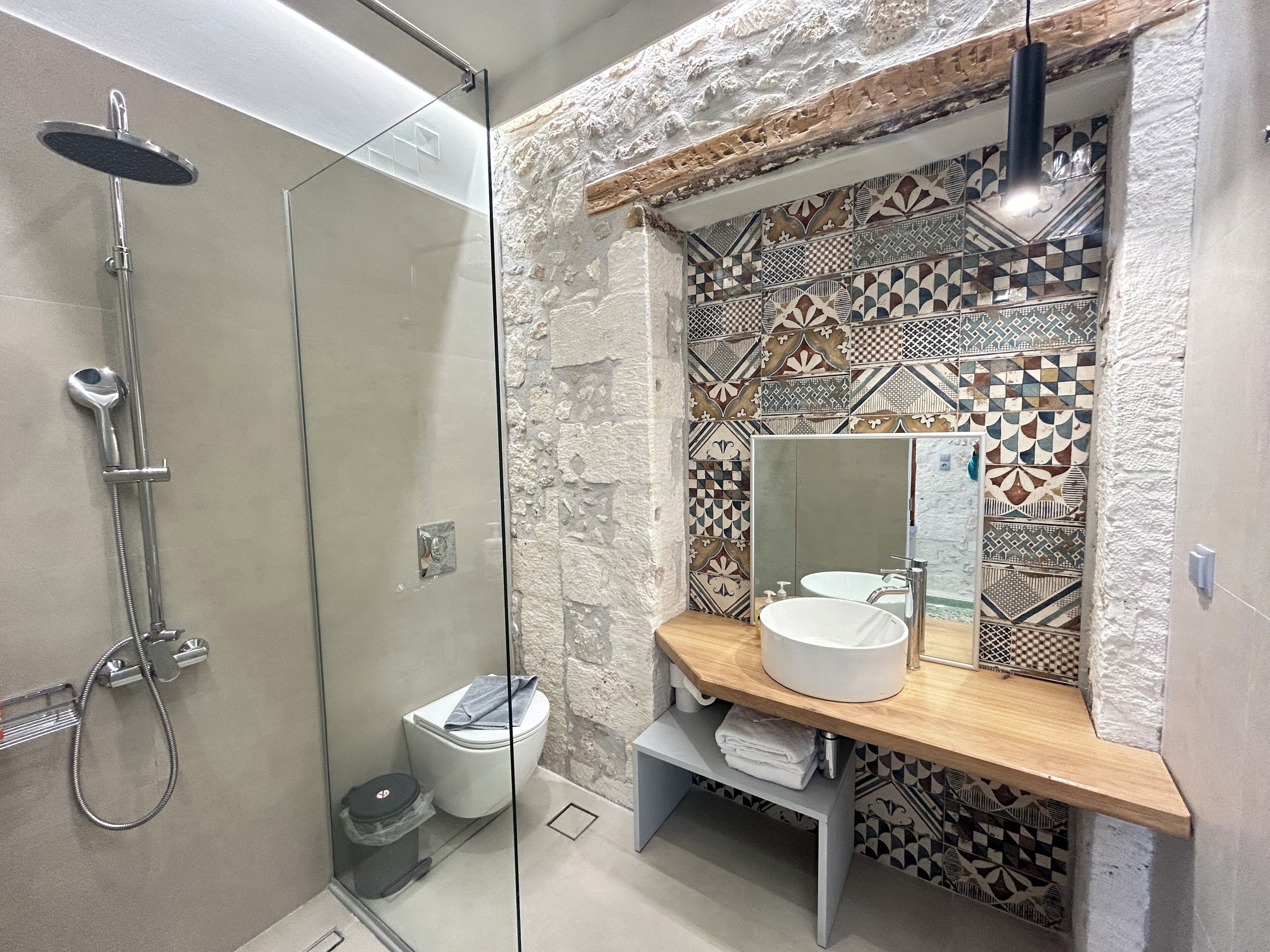 Un baño moderno con ducha envuelta en vidrio, un lavamanos blanco con espejo encima y paredes de azulejos estampados.