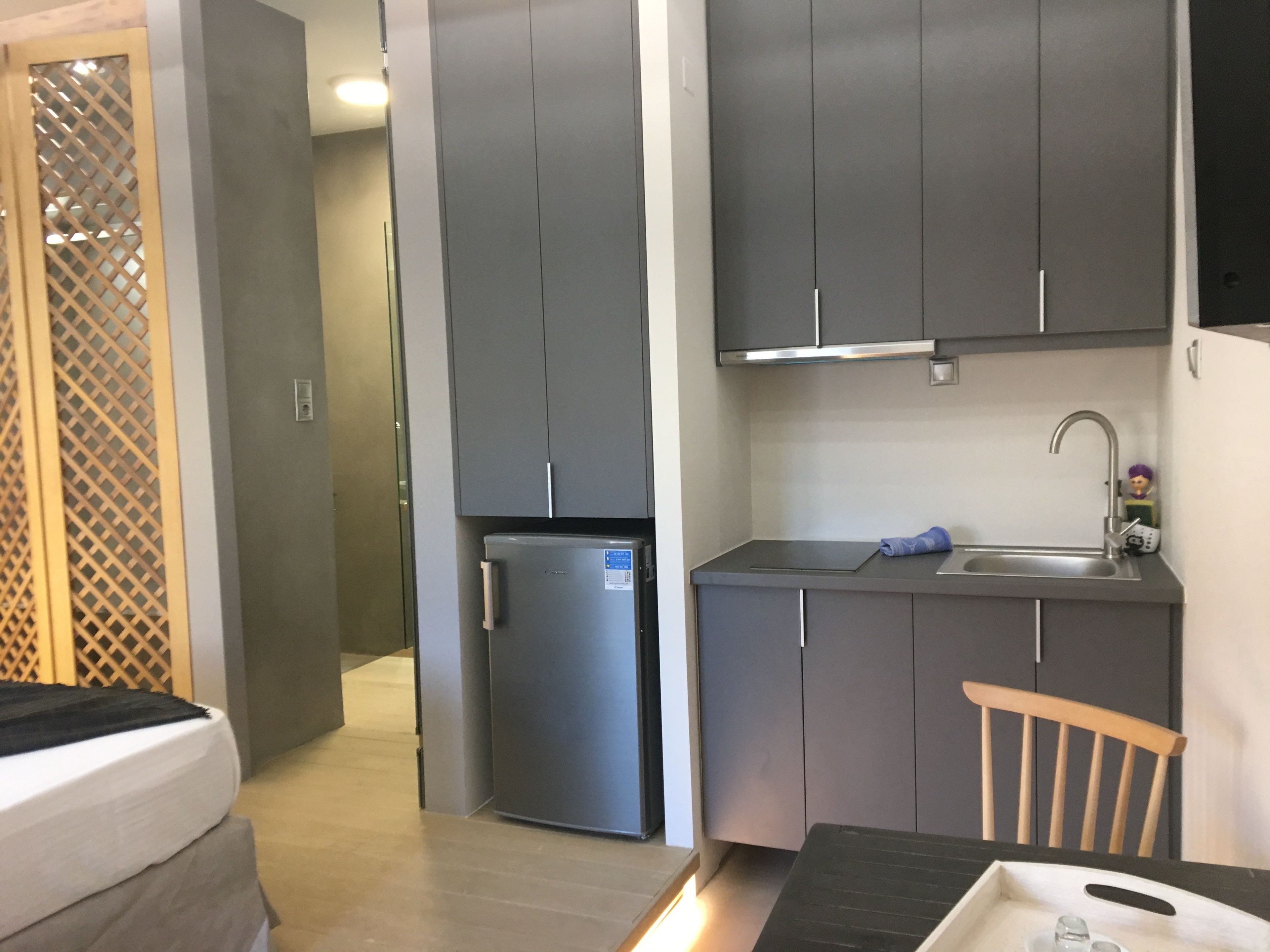 Eine kleine Küche mit grauen Schränken, einem Edelstahlkühlschrank, einer Spüle und einem Holzstuhl.