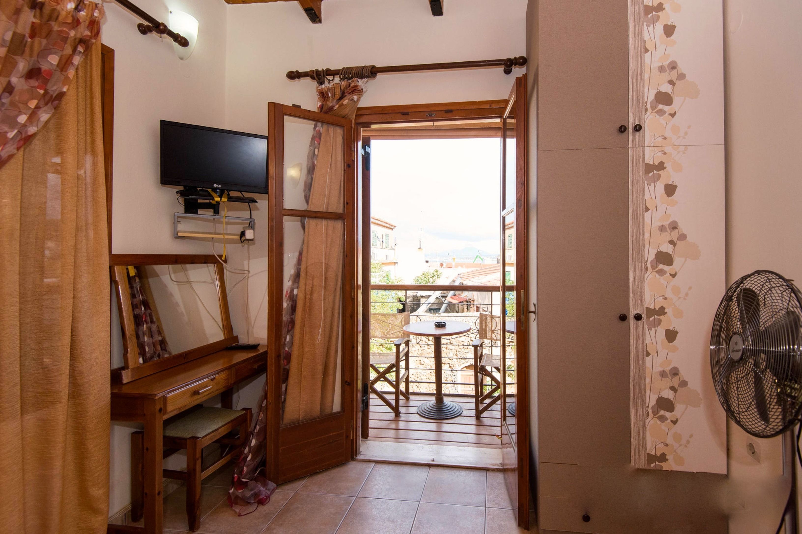 A room with a wooden desk, a television, a balcony with a table and chairs, and a fan.