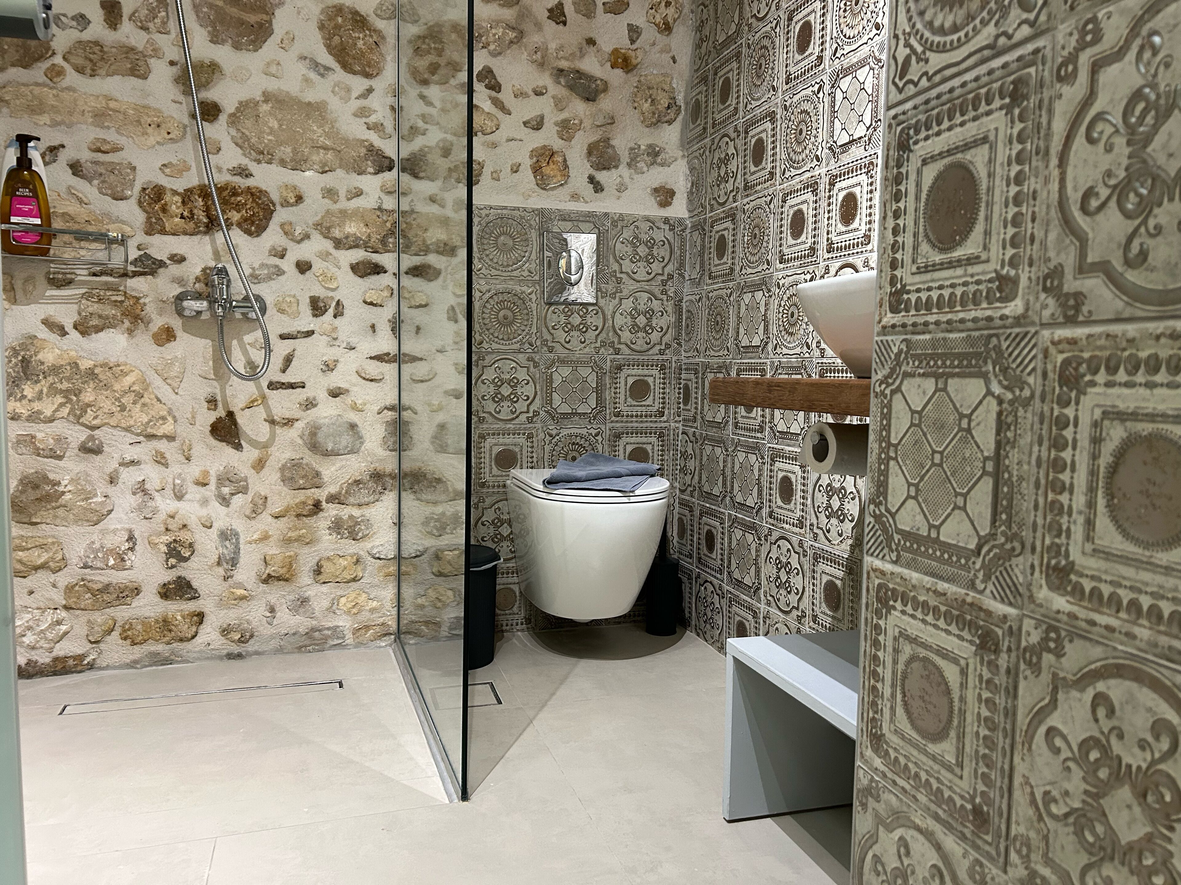 Un baño con pared de piedra, ducha, inodoro y baldosas estampadas.