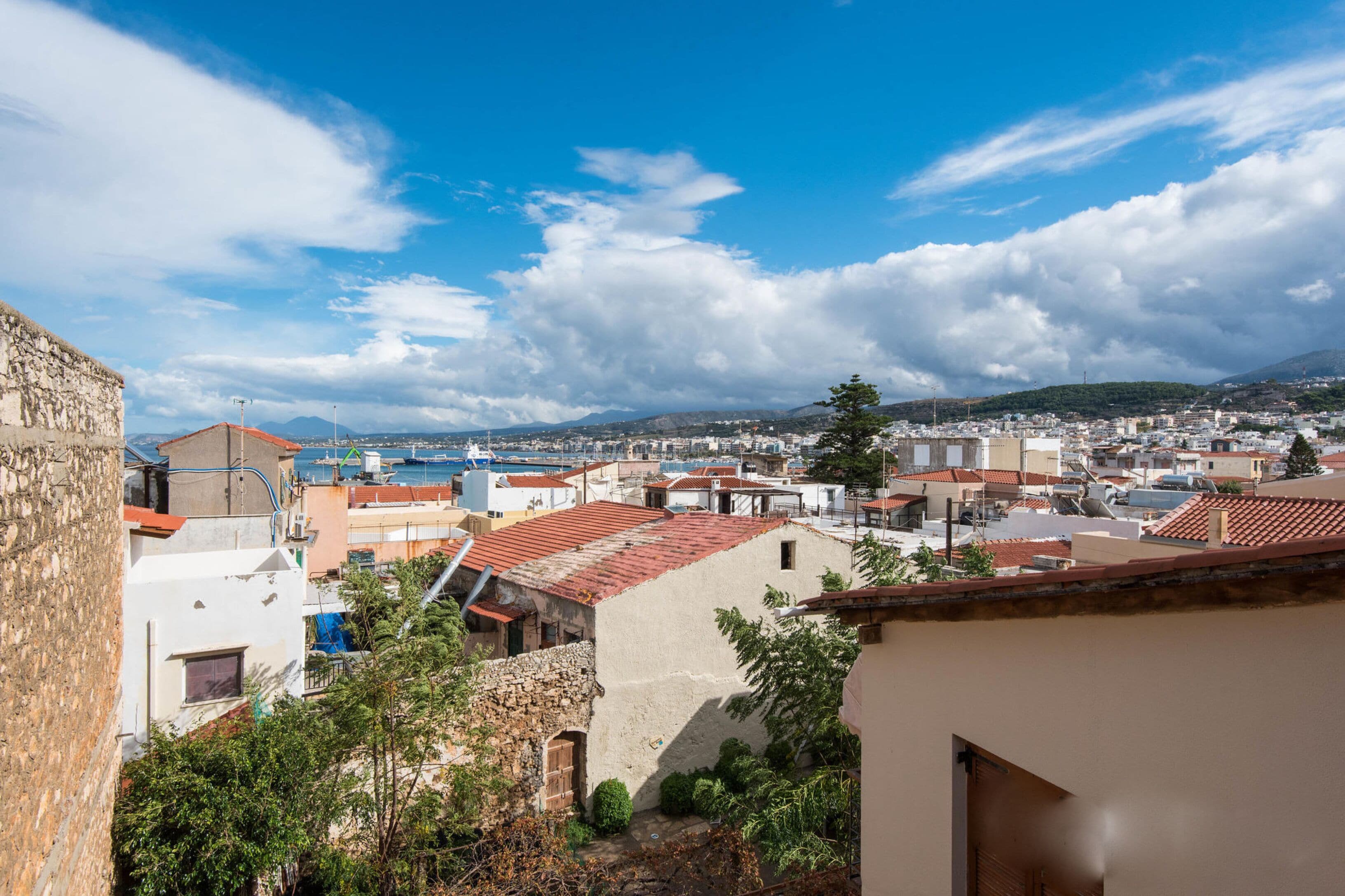 A coastal town with a mix of traditional and modern buildings, a clear sky, and a view of the sea and mountains in the distance.