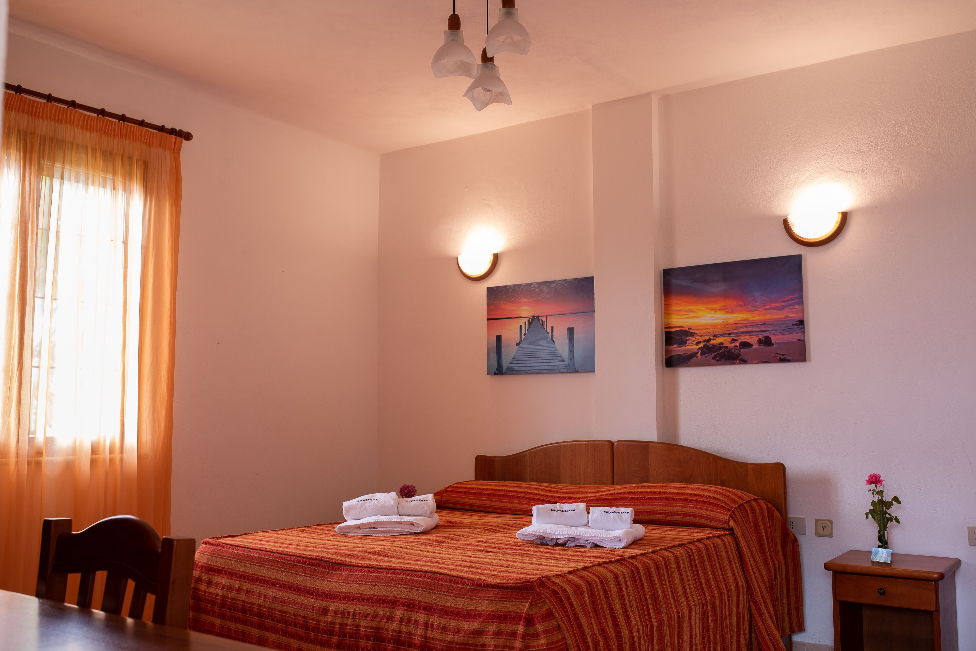 A hotel room with a wooden bed, two wall-mounted light fixtures, a painting of a sunset, and a small table with a vase of flowers.