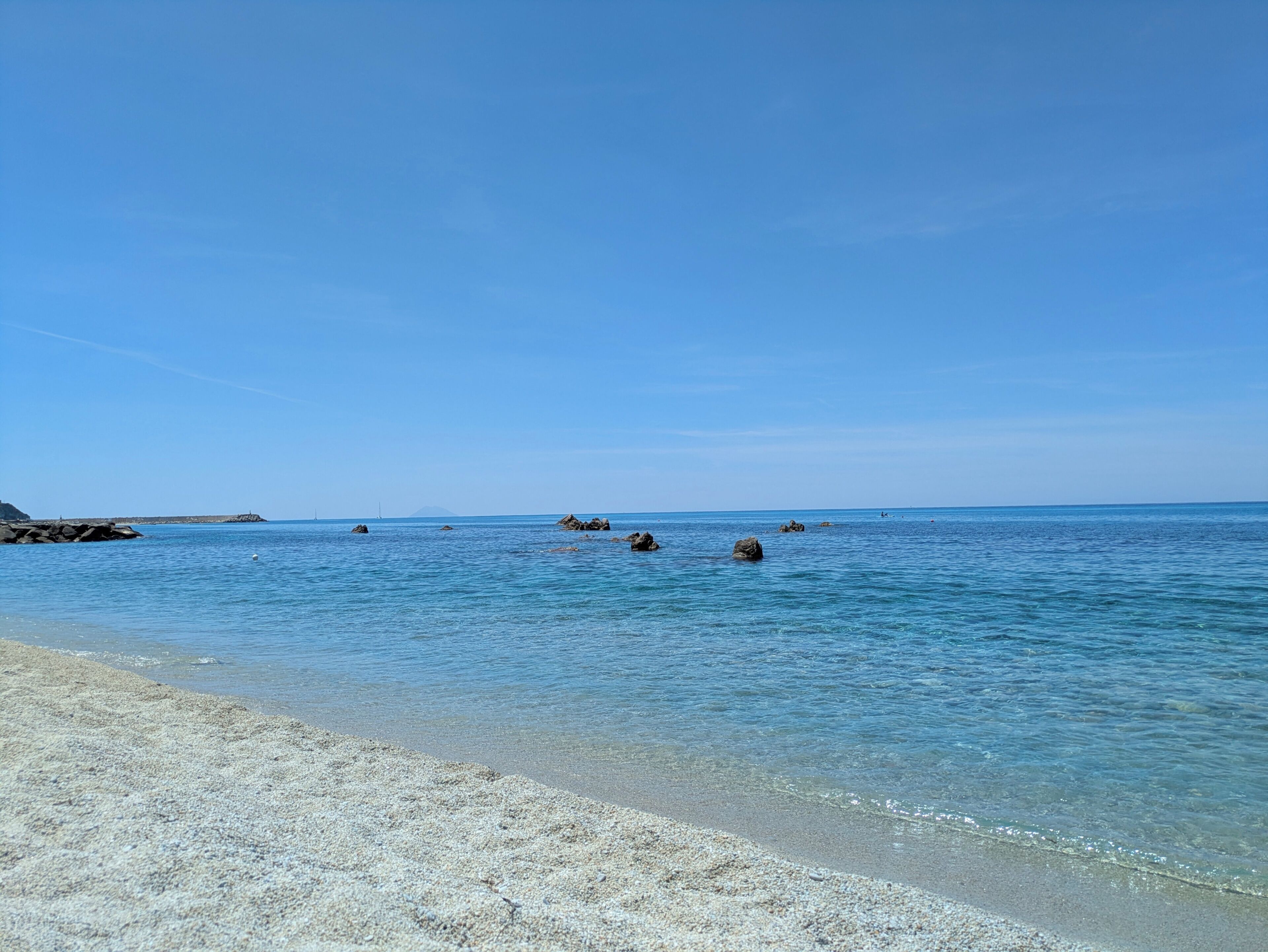 A beach with clear water, pebbly shore, and distant rocky outcrops.