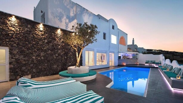 A modern pool area with a stone wall, a tree, and a white building in the background. - Gianna Suites (Santorini)