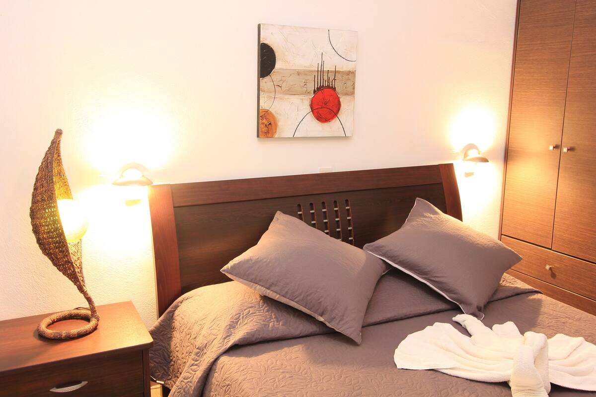 Una cama bien hecha con cabecera, dos almohadas, una manta y una bata. Hay una lámpara en una mesita de noche y una obra de arte en la pared sobre la cama.