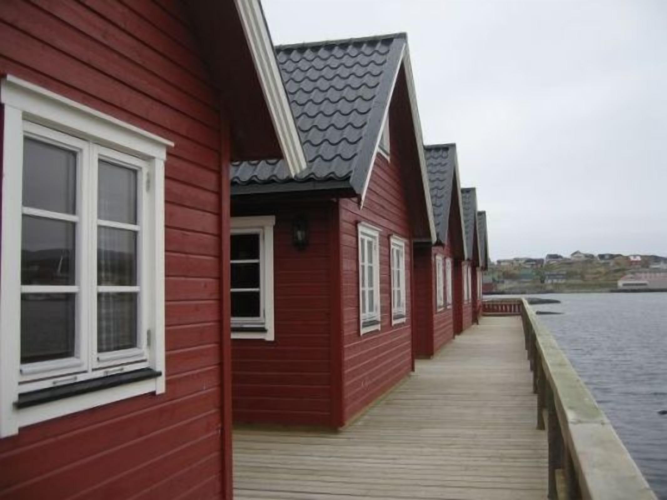 Une rangée de bâtiments en bois rouge, dotés de fenêtres blanches et d’une terrasse en bois qui s’étend au-dessus d’une étendue d’eau.