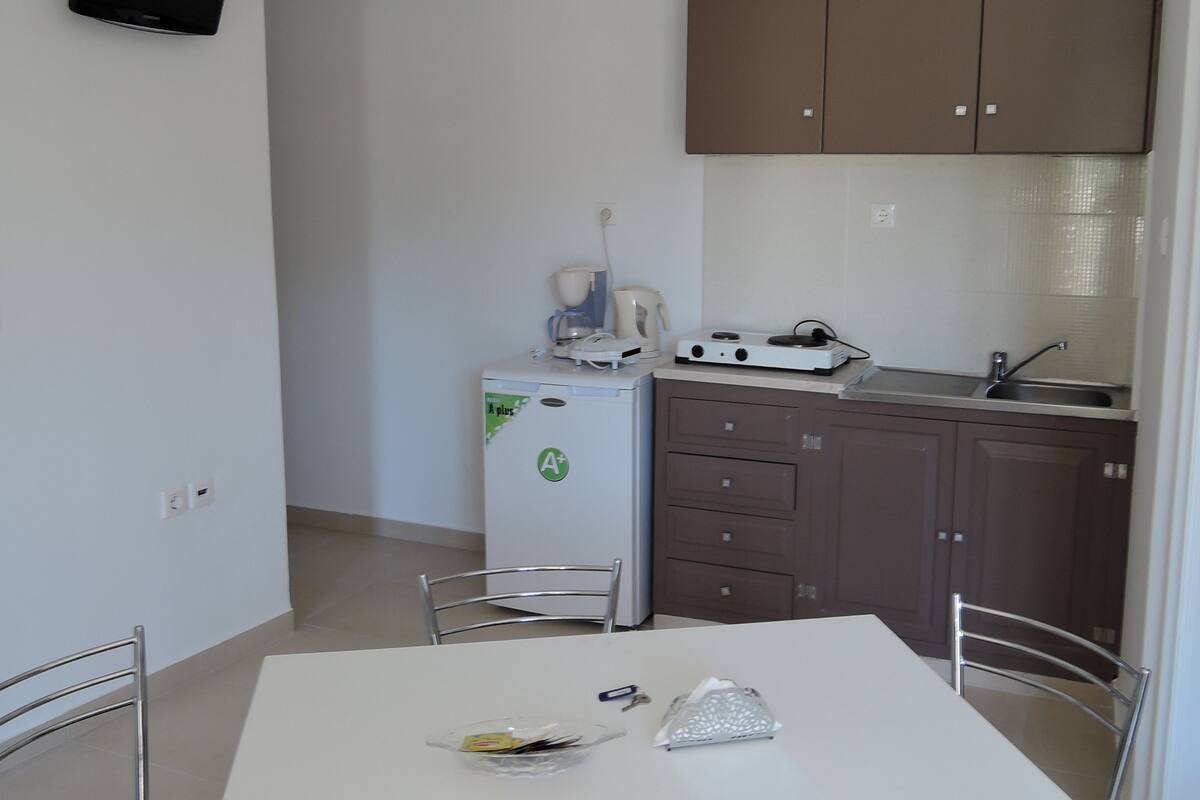 Une cuisine compacte équipée d’un réfrigérateur, d’une machine à café et d’un four à micro-ondes. On y trouve une table à manger avec des chaises, ainsi qu’un téléviseur fixé au mur.