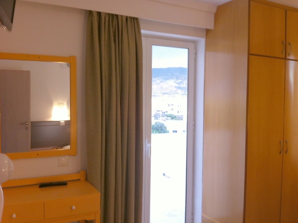 Quarto de hotel com guarda-roupa de madeira, espelho, mesinha com abajur e vista para prédios através de uma janela com cortinas.