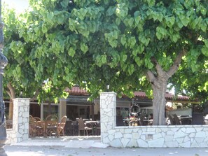 An outdoor seating area with stone pillars, wooden chairs, and tables, surrounded by lush green foliage. - Hotel Panorama (Aigialeia)