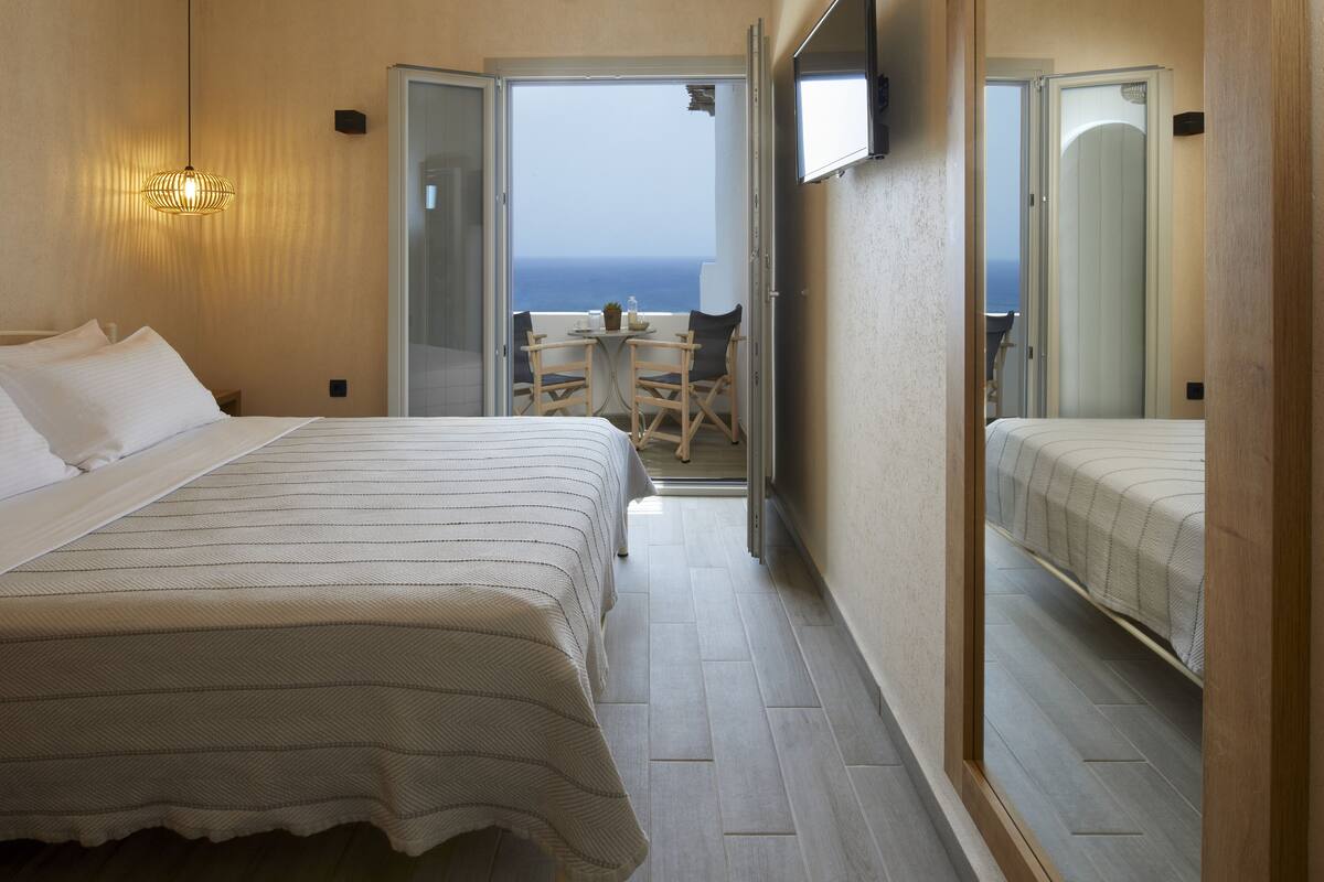 A hotel room with a bed, a small table with chairs, a TV, and a balcony with a view of the ocean.
