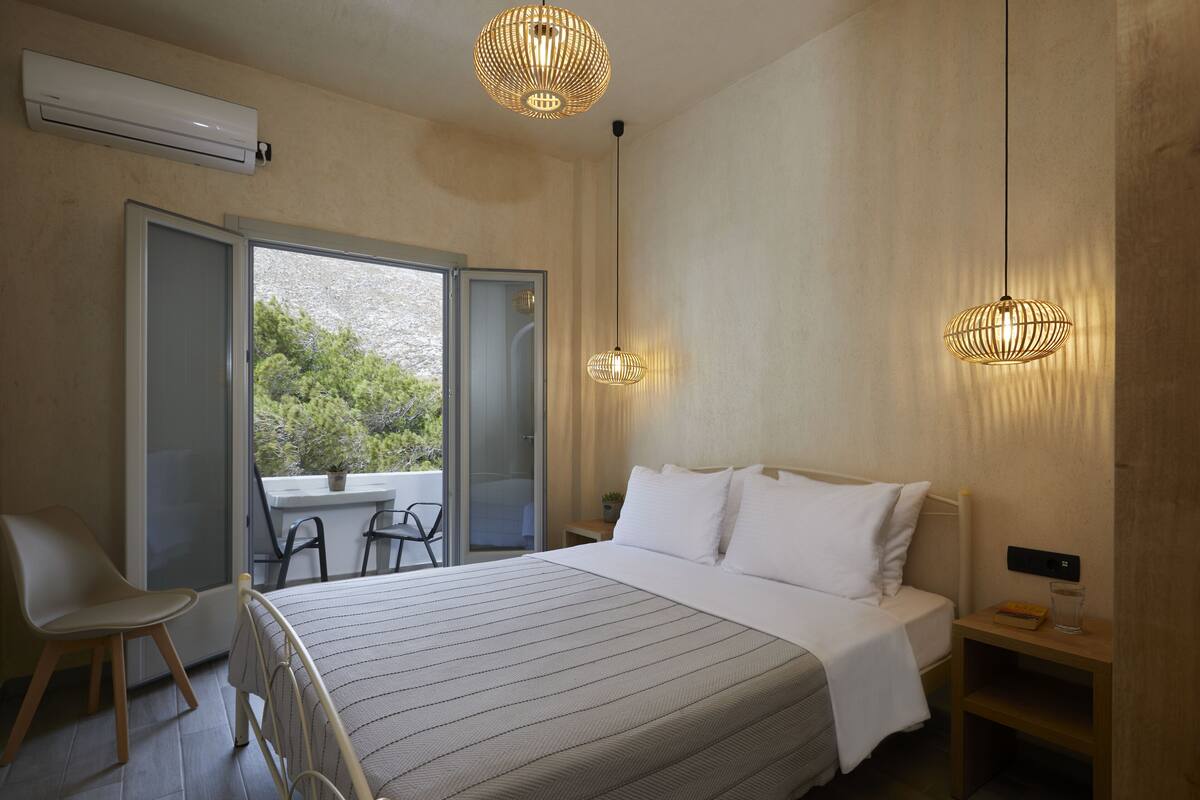 A modern bedroom with a large bed, two pendant lights, a bedside table, and a view of greenery outside.