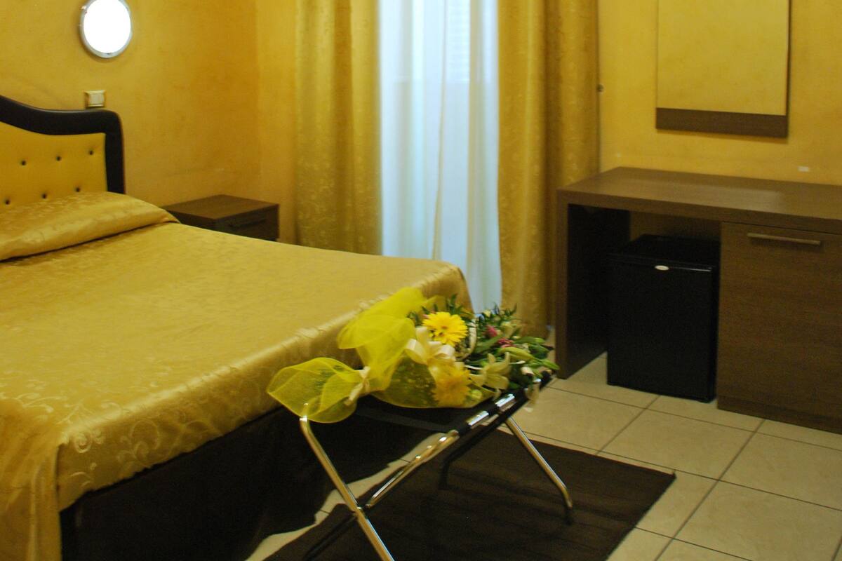 A hotel room with a bed, a bedside table, an air conditioning unit, a mirror, and a window with curtains.