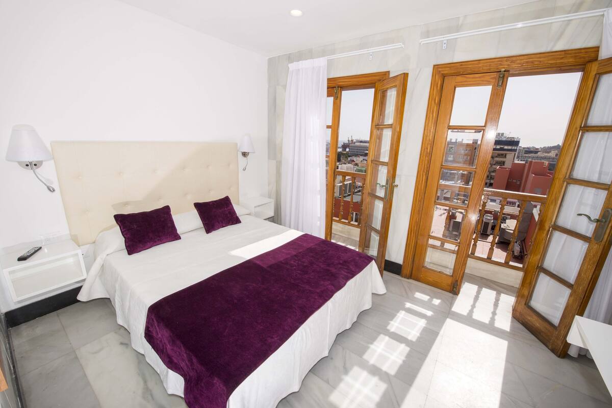 A hotel room with a bed, a nightstand, a lamp, and a view of the city through a glass door.
