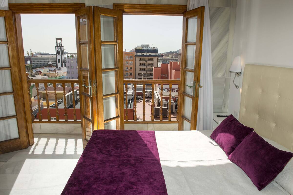 A room with a bed, a view of buildings through open doors, and a balcony railing.
