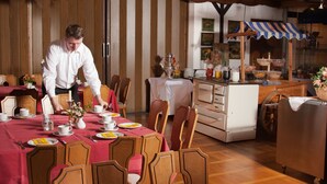 Comedor con sillas de madera y una mesa dispuesta para una comida, zona de cocina con bufé y una persona sirviendo comida.