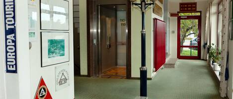 A hallway with a green patterned carpet, an elevator, a red door, and a wall with framed pictures and signs.