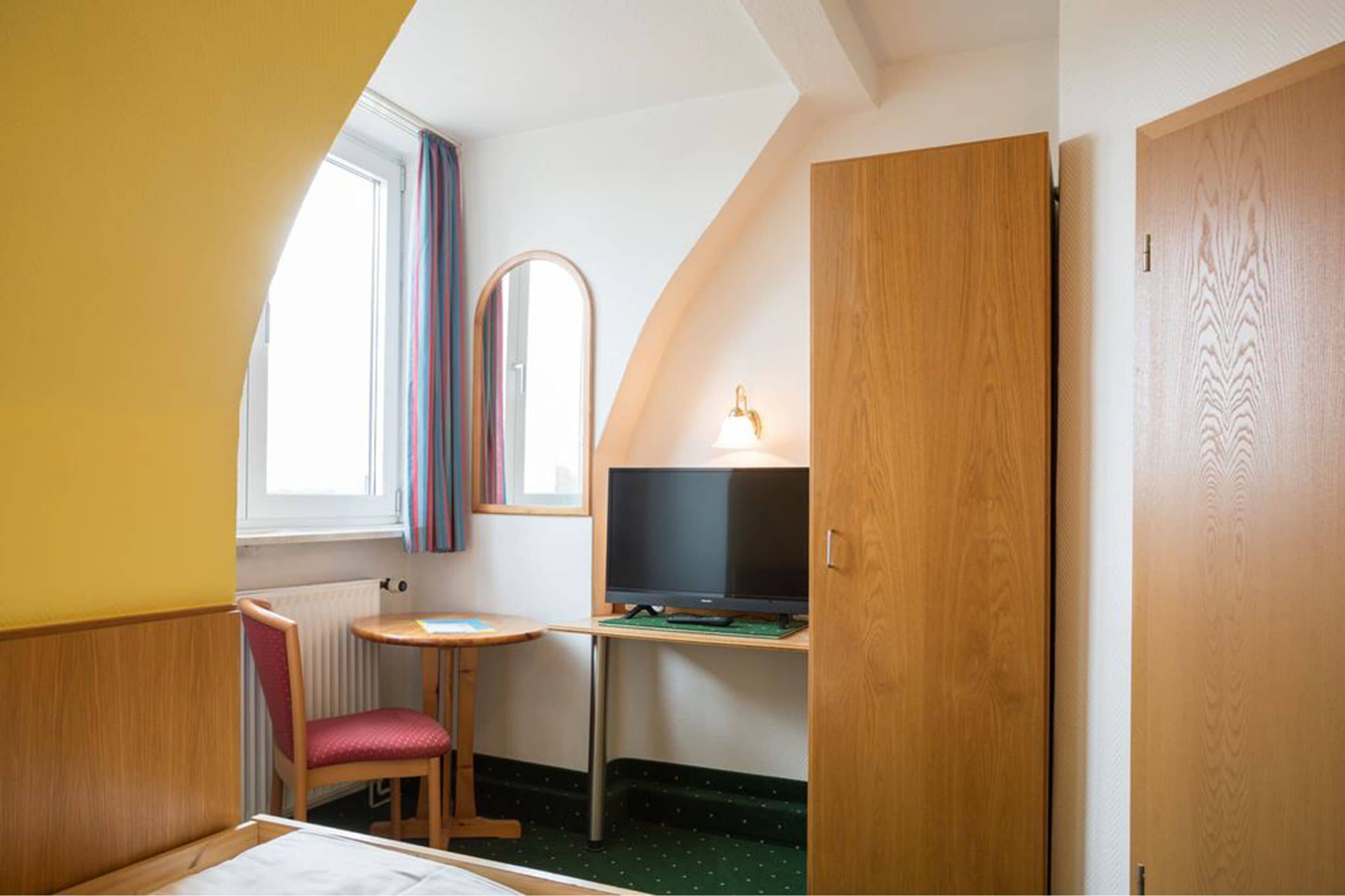 A small, cozy room with a bed, a desk with a computer, a chair, a window with curtains, and a wooden door.
