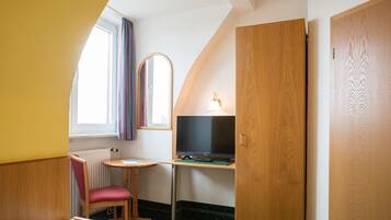 A small, cozy room with a bed, a desk with a computer, a chair, a window with curtains, and a wooden door.