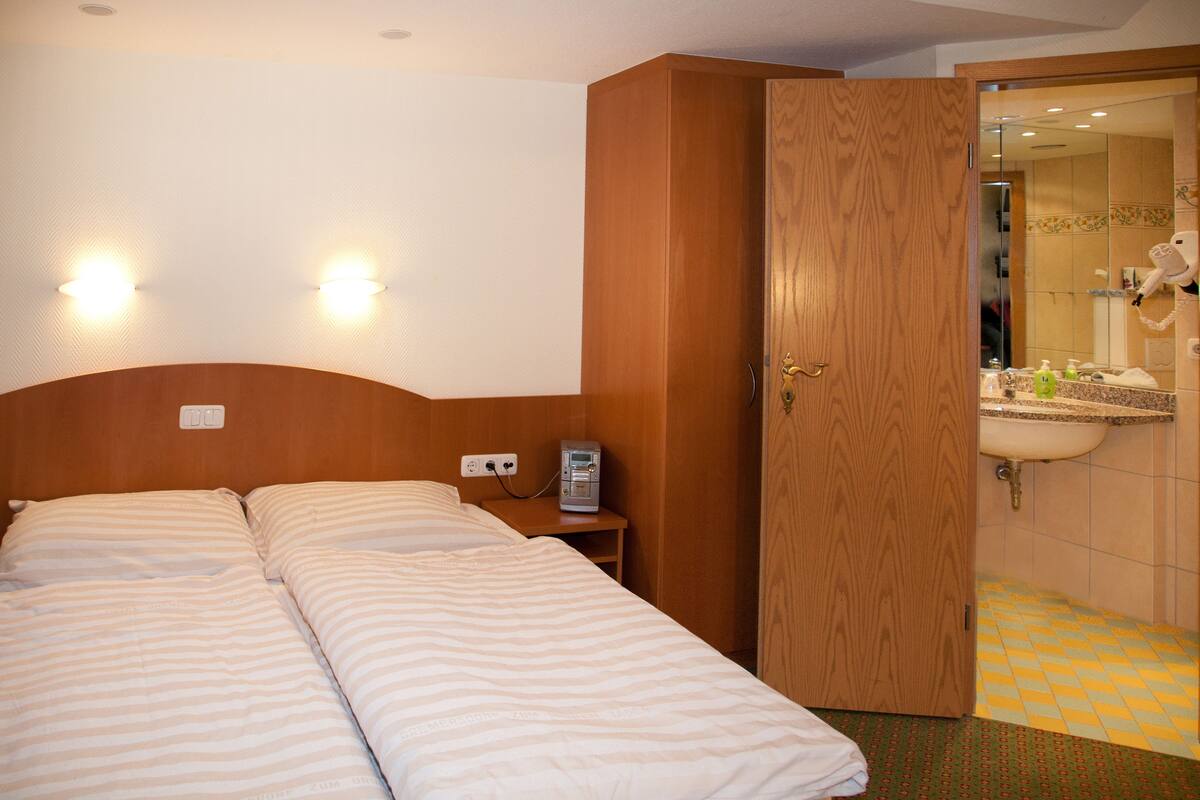 A hotel room with two beds, a wooden wardrobe, a bedside table with a phone, and a bathroom with a sink and mirror.