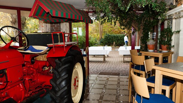Un tractor rojo estacionado bajo un toldo a rayas, una mesa de madera con sillas y un mantel blanco al fondo.