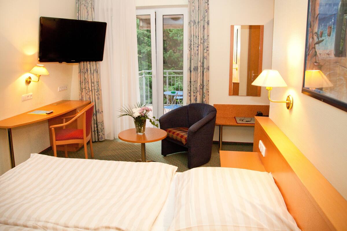 A hotel room with a bed, a desk, a chair, a TV, and a balcony with a table and chairs.