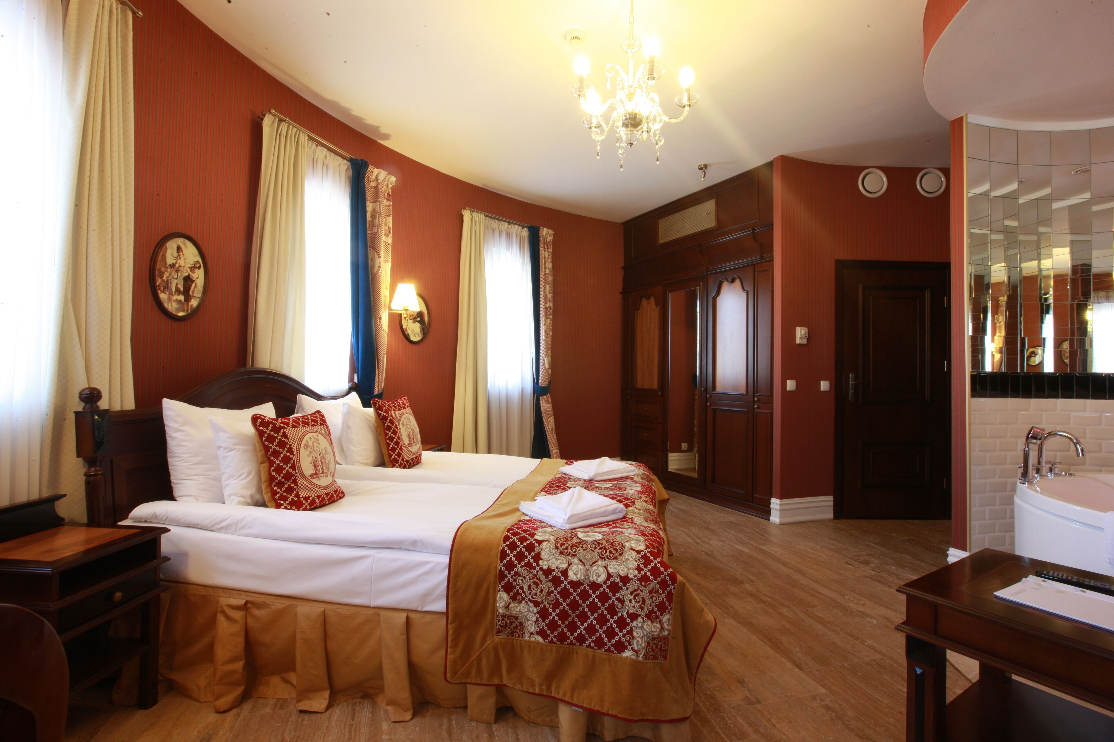 Ein Hotelzimmer mit einem großen Bett, einem Schreibtisch, einem Sessel, einer Kandelaberleuchte, einem Badezimmer mit Waschbecken und Spiegel sowie einer Tür zu einem weiteren Raum.
