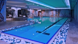 En stor indendørs swimmingpool med krystalklart vand, omgivet af hvide og blå sten, samt med liggestole og omklædningsrum.