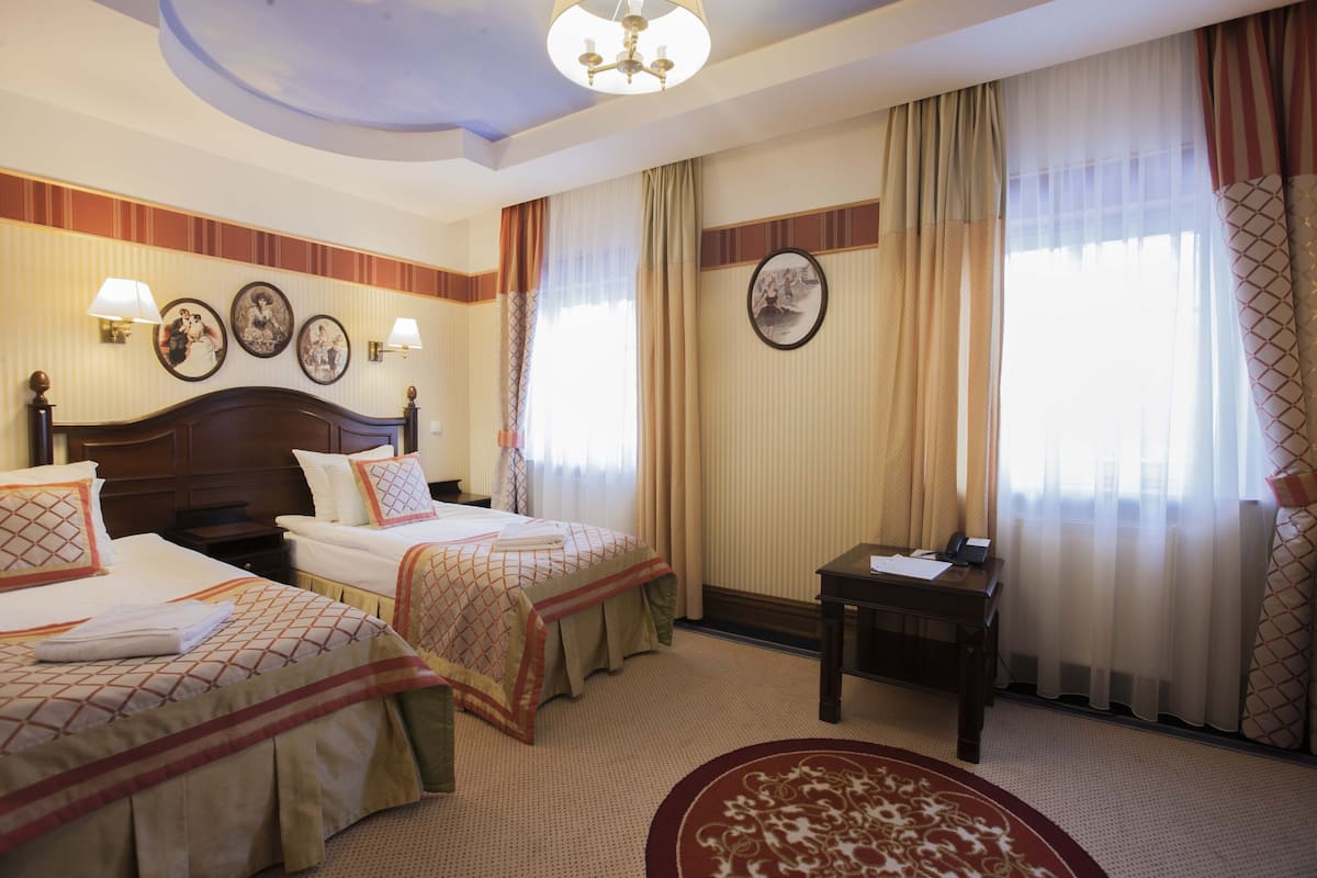 Habitación de hotel con dos camas, un escritorio y una silla. Hay lámparas de pared, un candelabro y cortinas.