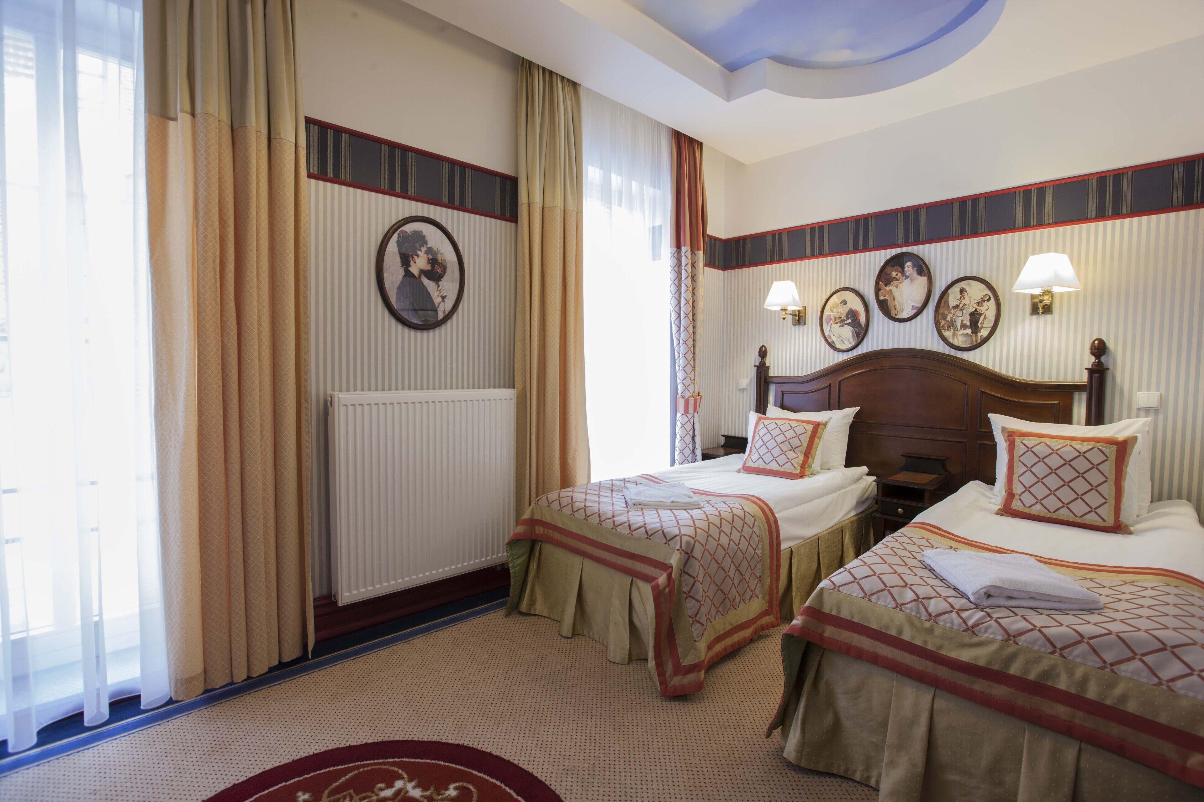 A hotel room with two beds, a large window with curtains, a radiator, and wall-mounted lamps.