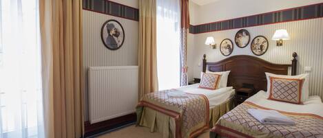 A hotel room with two beds, a large window with curtains, a radiator, and wall-mounted lamps.