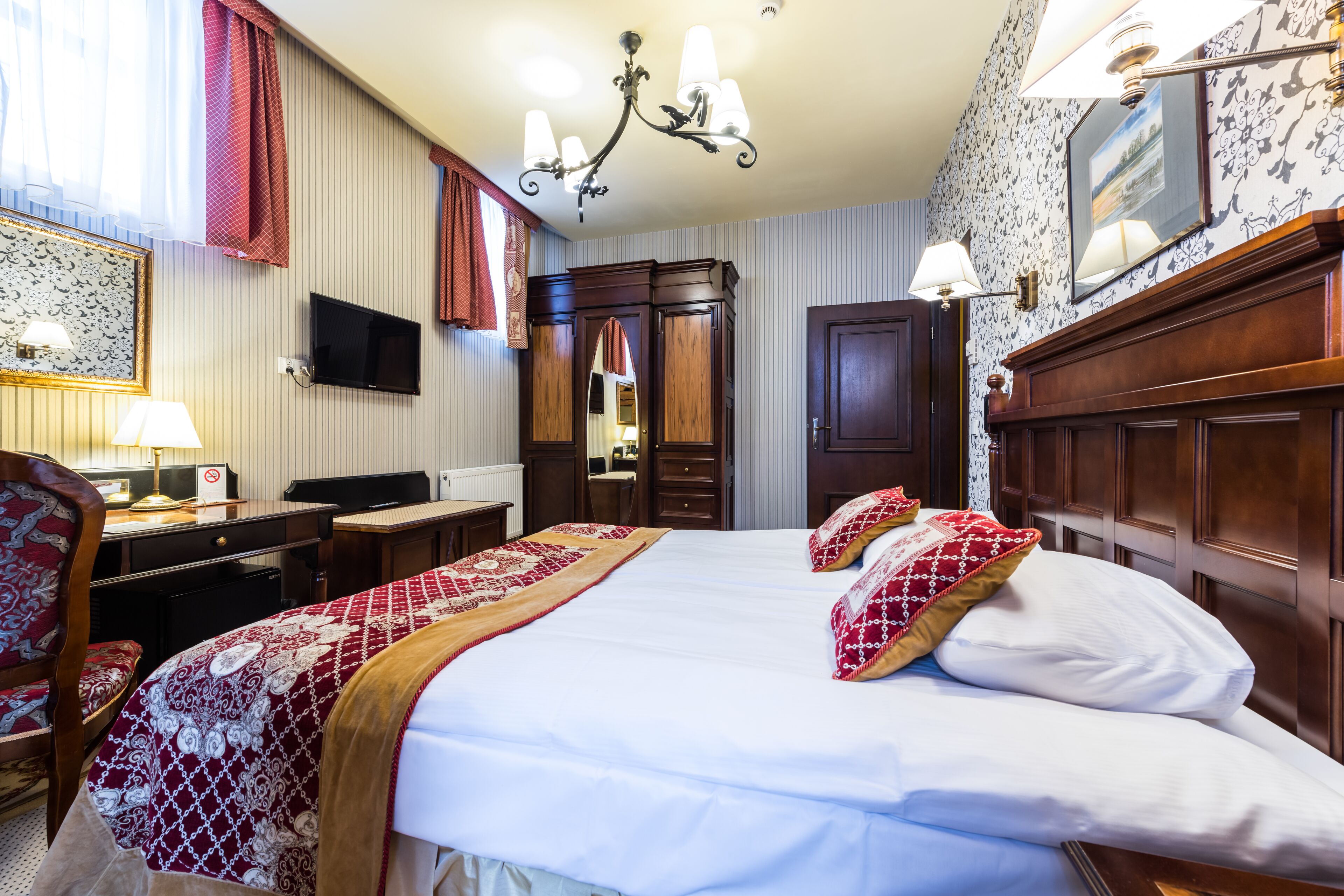 A hotel room with a large bed, a desk with a computer, a TV, and a chandelier.