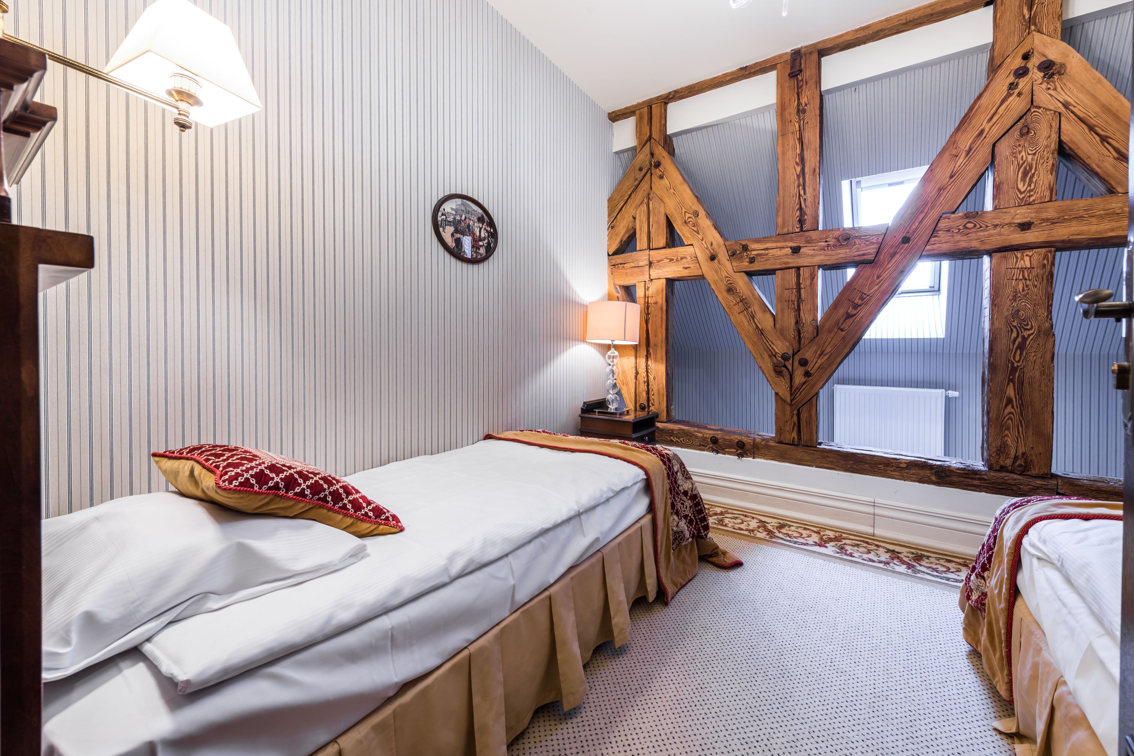 A room with two beds, striped wallpaper, a wooden beam ceiling, a window, and a lamp on a nightstand.
