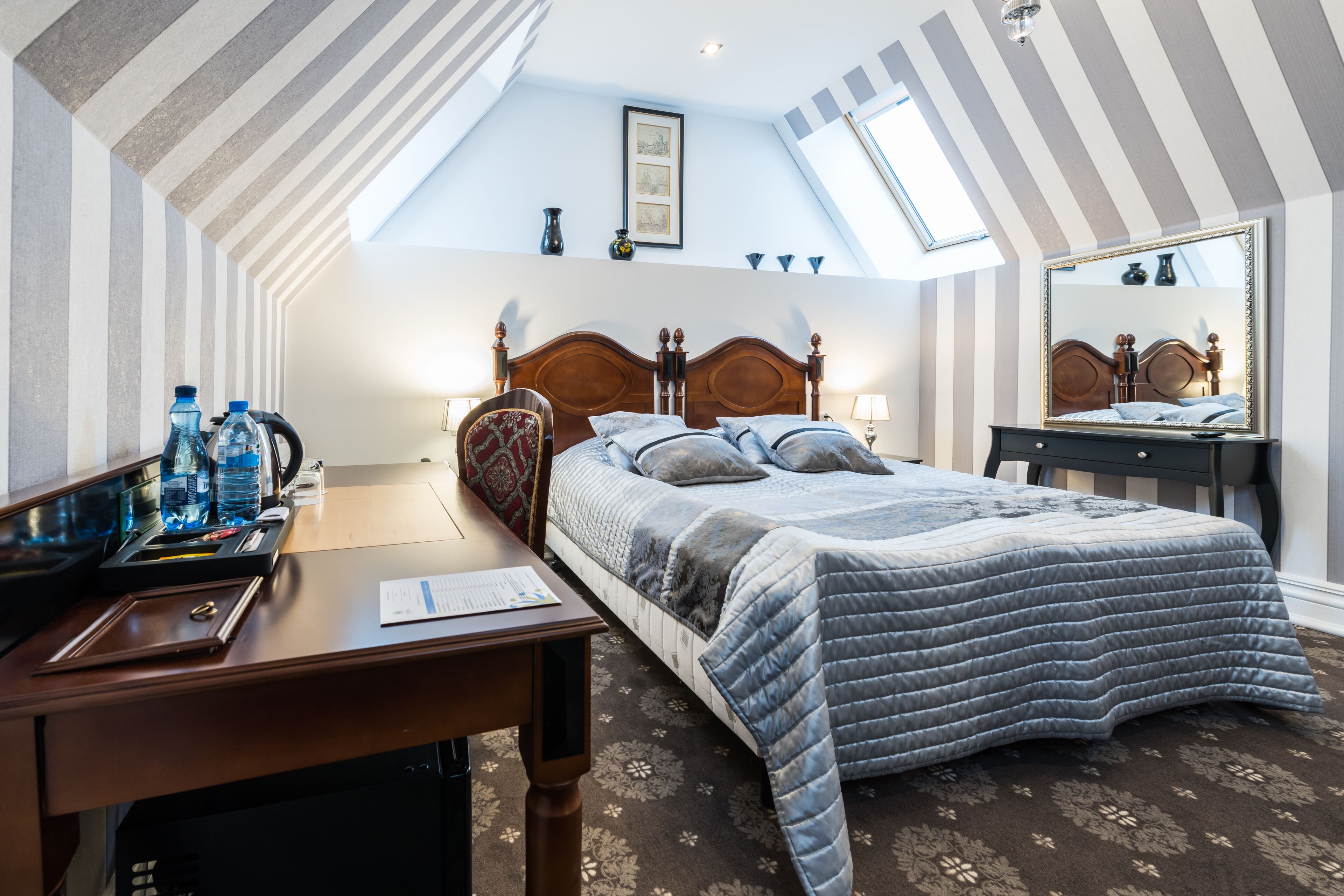 A bedroom with a sloped ceiling, a bed with a quilted cover, a wooden desk with a chair, and a mirror on the wall.