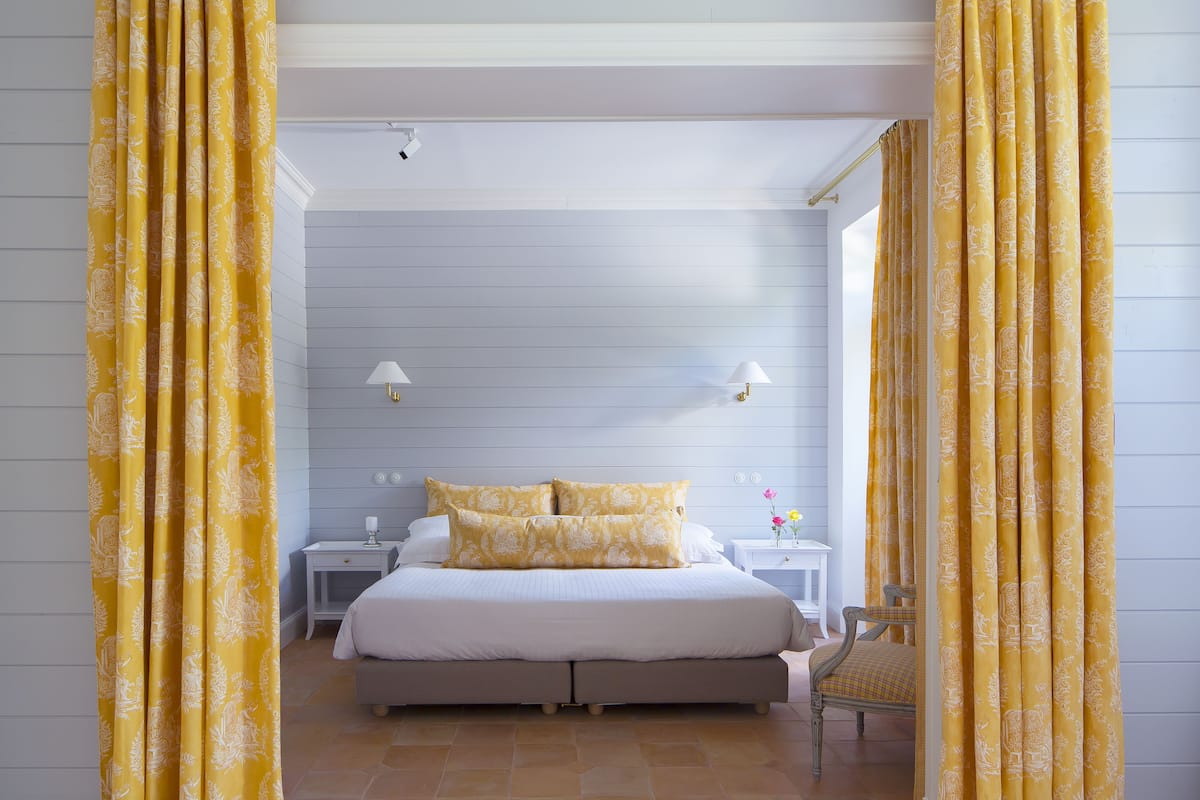 Ein Schlafzimmer mit einem Bett, zwei Nachttischen, einem Sessel und gelben Vorhängen.
