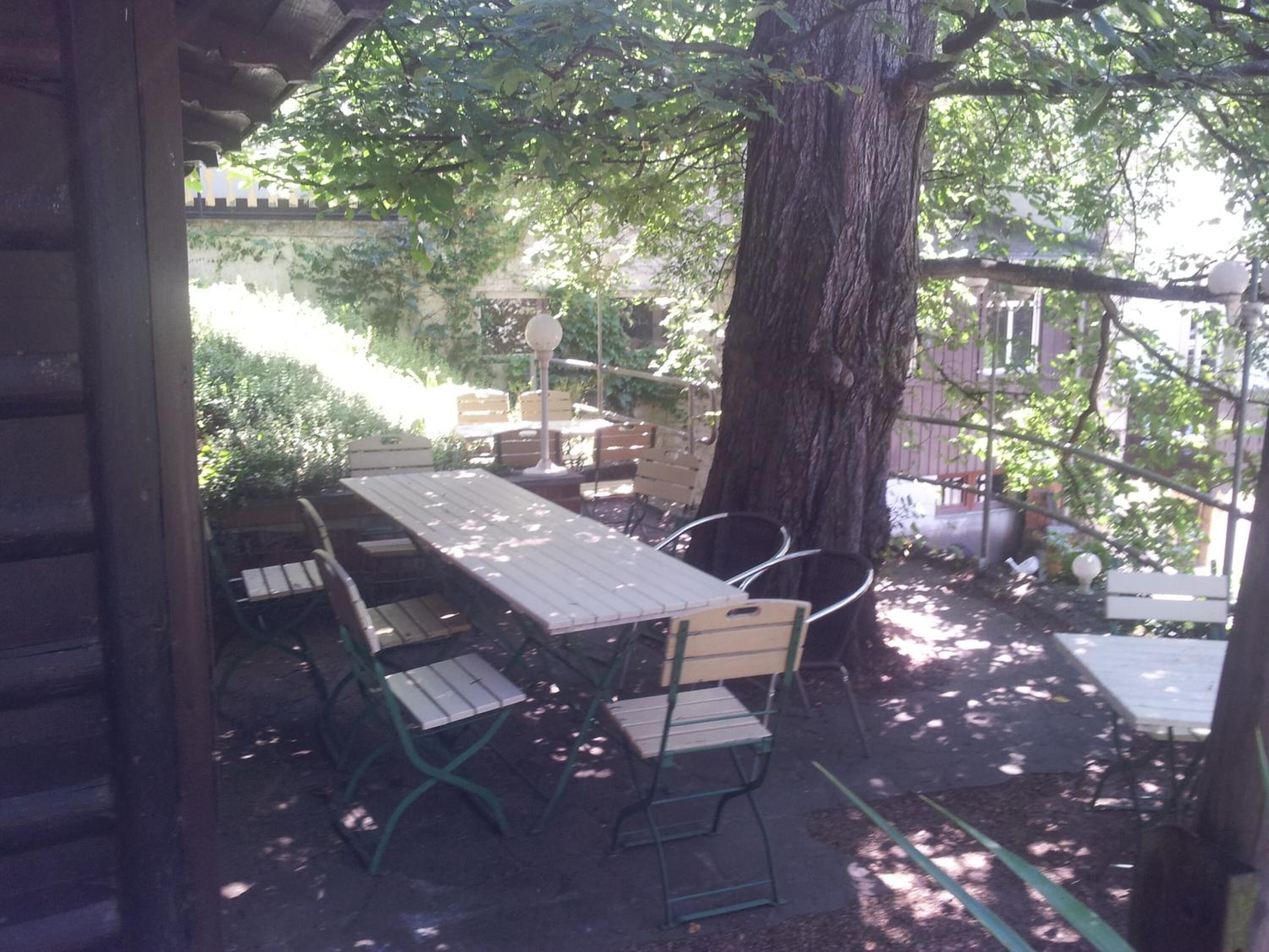An outdoor patio with a wooden table and chairs, surrounded by trees and a brick wall.