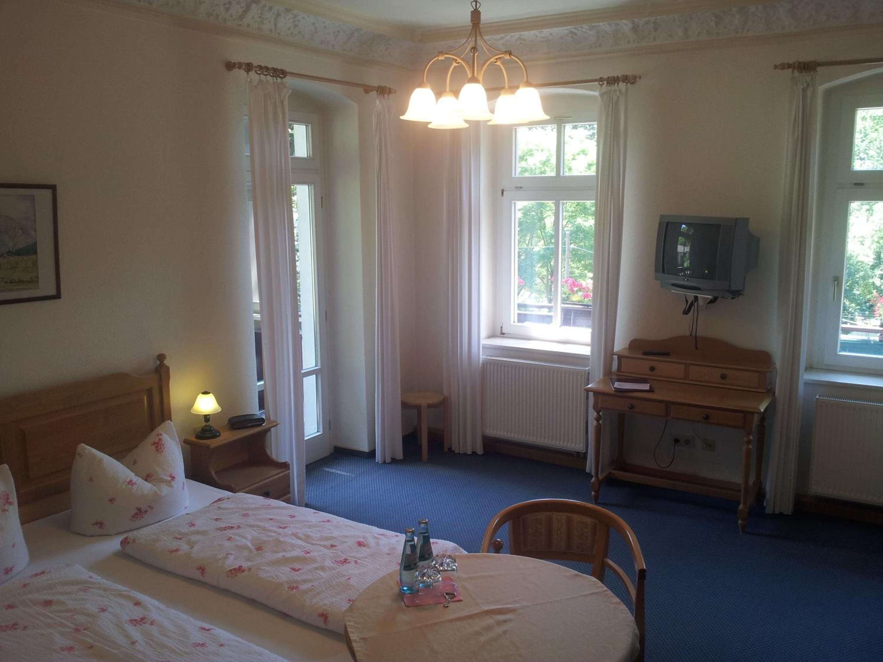 Een hotelkamer met een bed, nachtkastje, lamp, dressoir, televisie en ramen.