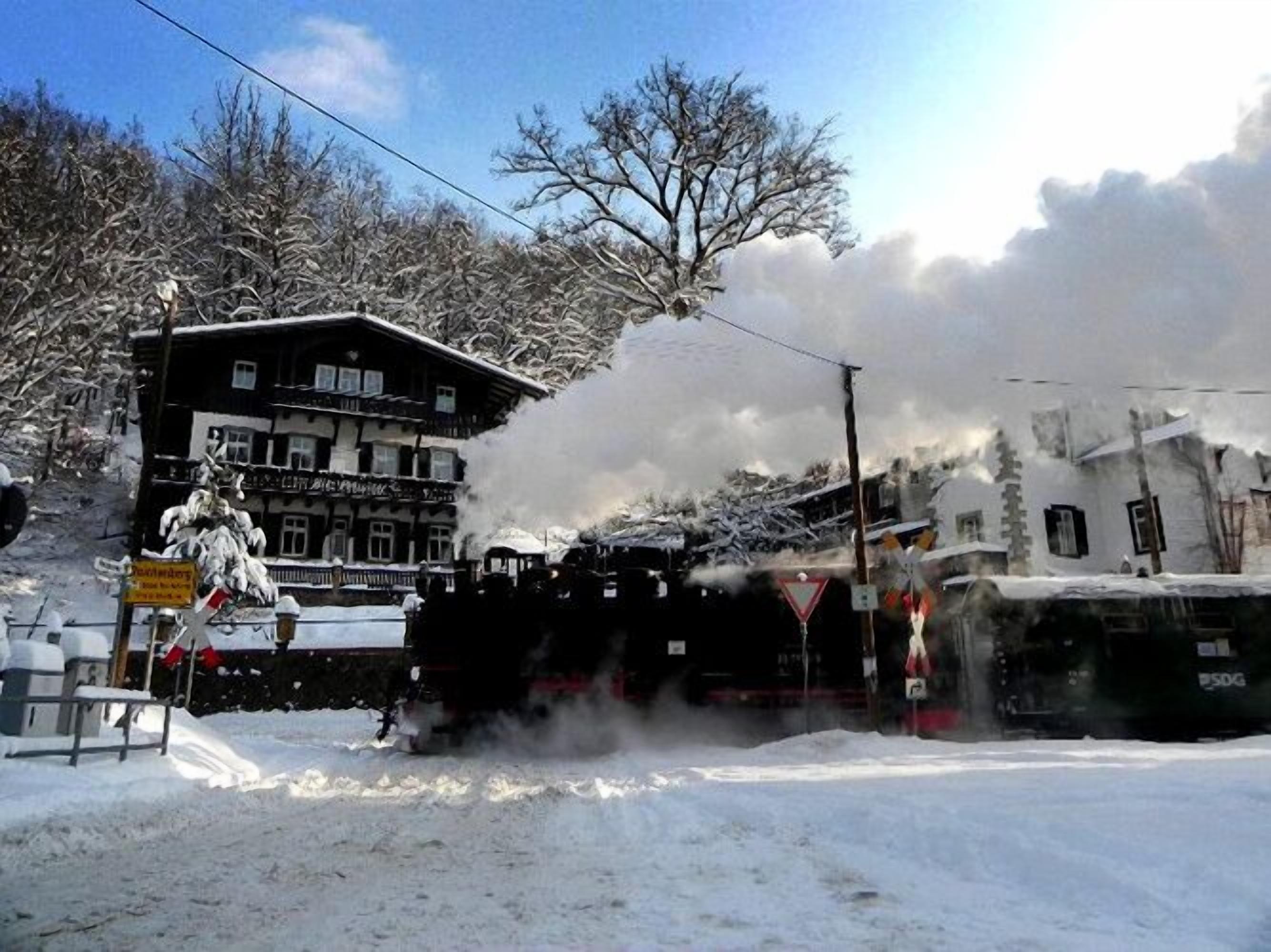 Una estación de tren con una locomotora de vapor que expulsa humo, rodeada de edificios y árboles cubiertos de nieve.