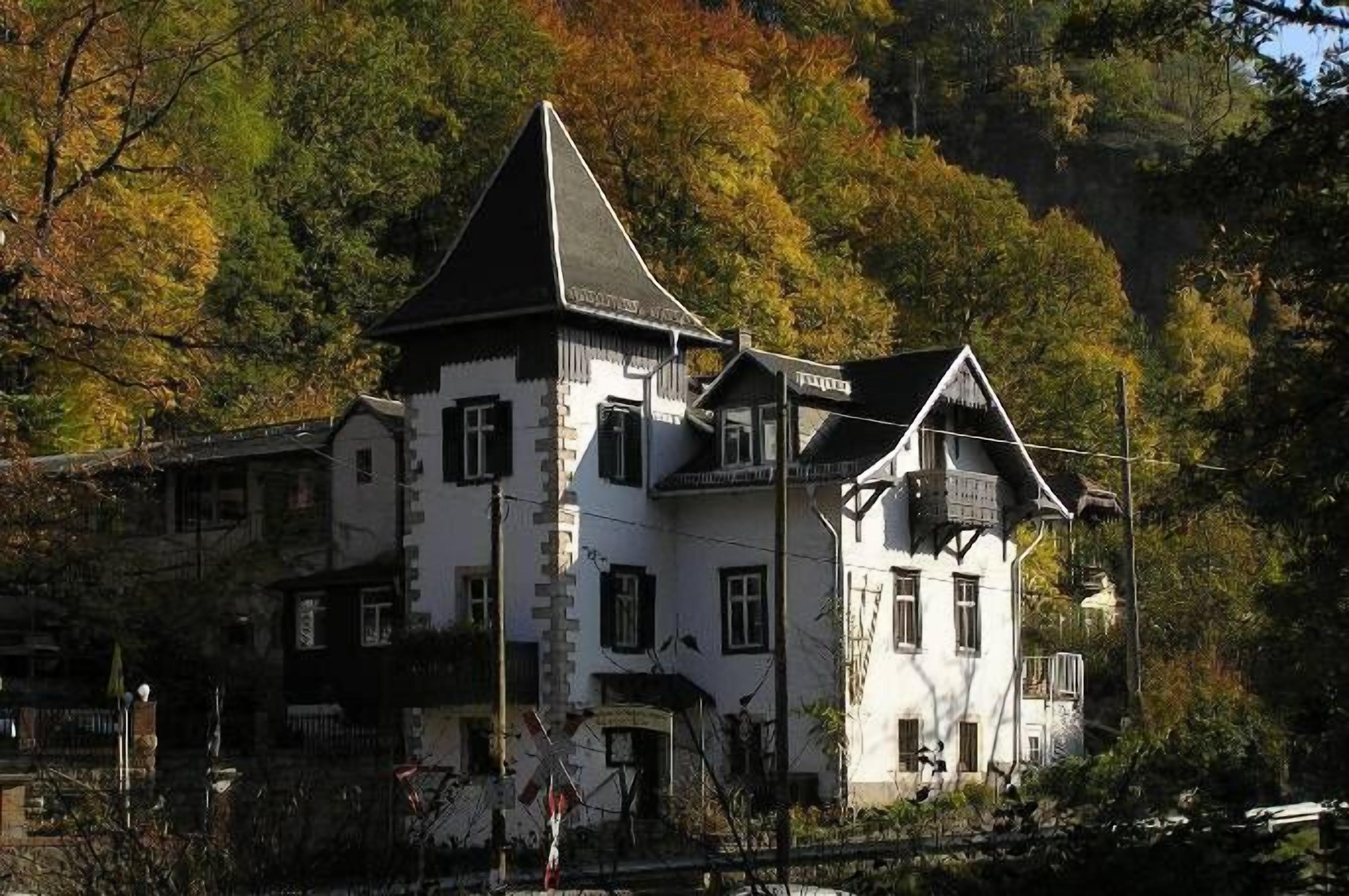 Un edificio blanco con múltiples ventanas y un techo empinado, rodeado de árboles y un terreno montañoso rocoso.