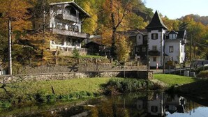 Un edificio de estilo europeo tradicional con un puente de piedra sobre un río tranquilo, rodeado de follaje otoñal.
