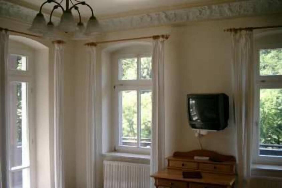 A room with a wooden desk, a television mounted on the wall, and a window with curtains.