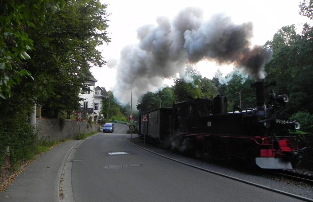 Eine Dampflokomotive stößt dichten Rauch auf Gleisen neben einer Straße mit einem Auto aus.