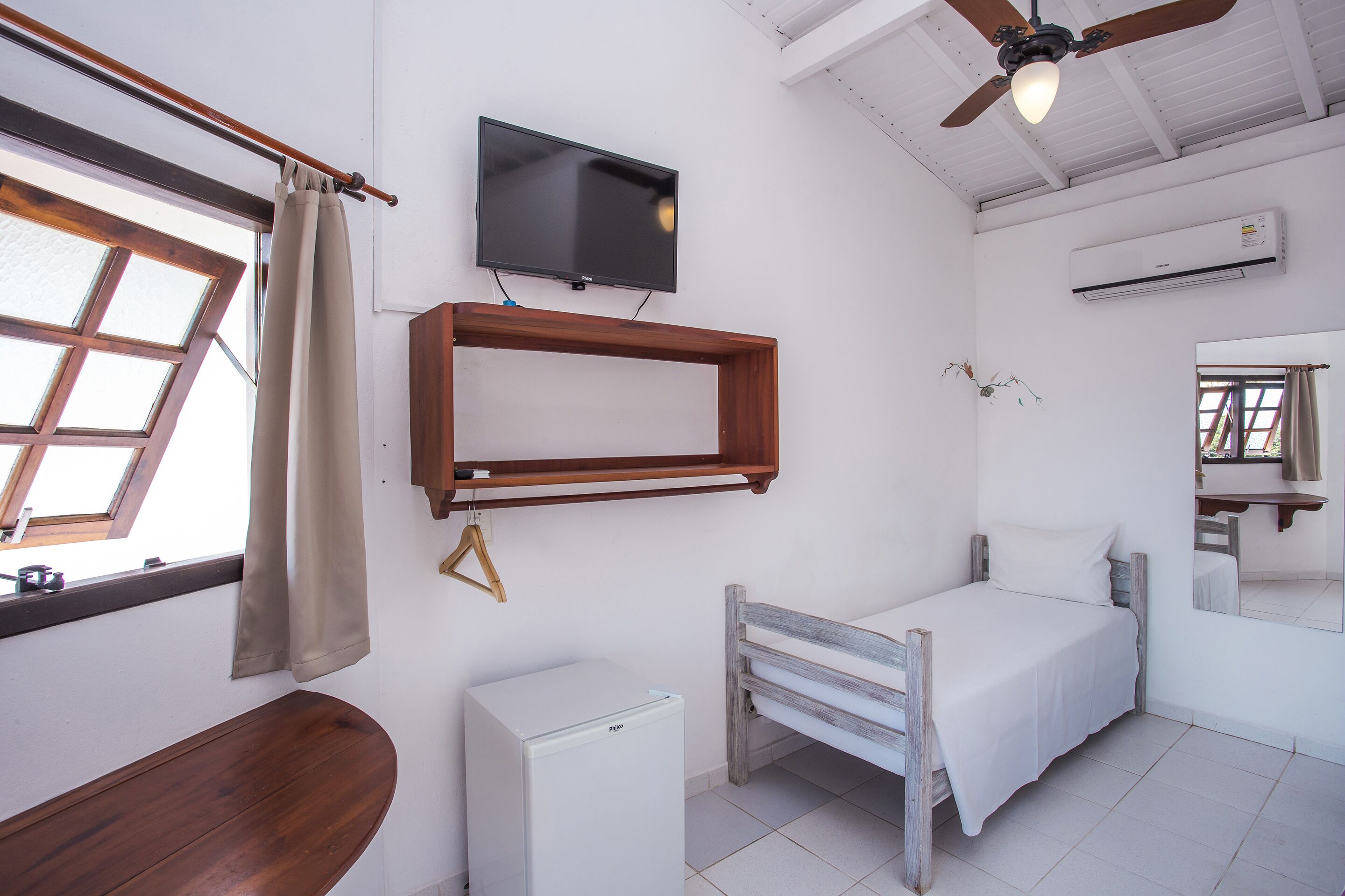 A compact room with a bed, a small white refrigerator, a mounted TV, and a ceiling fan.