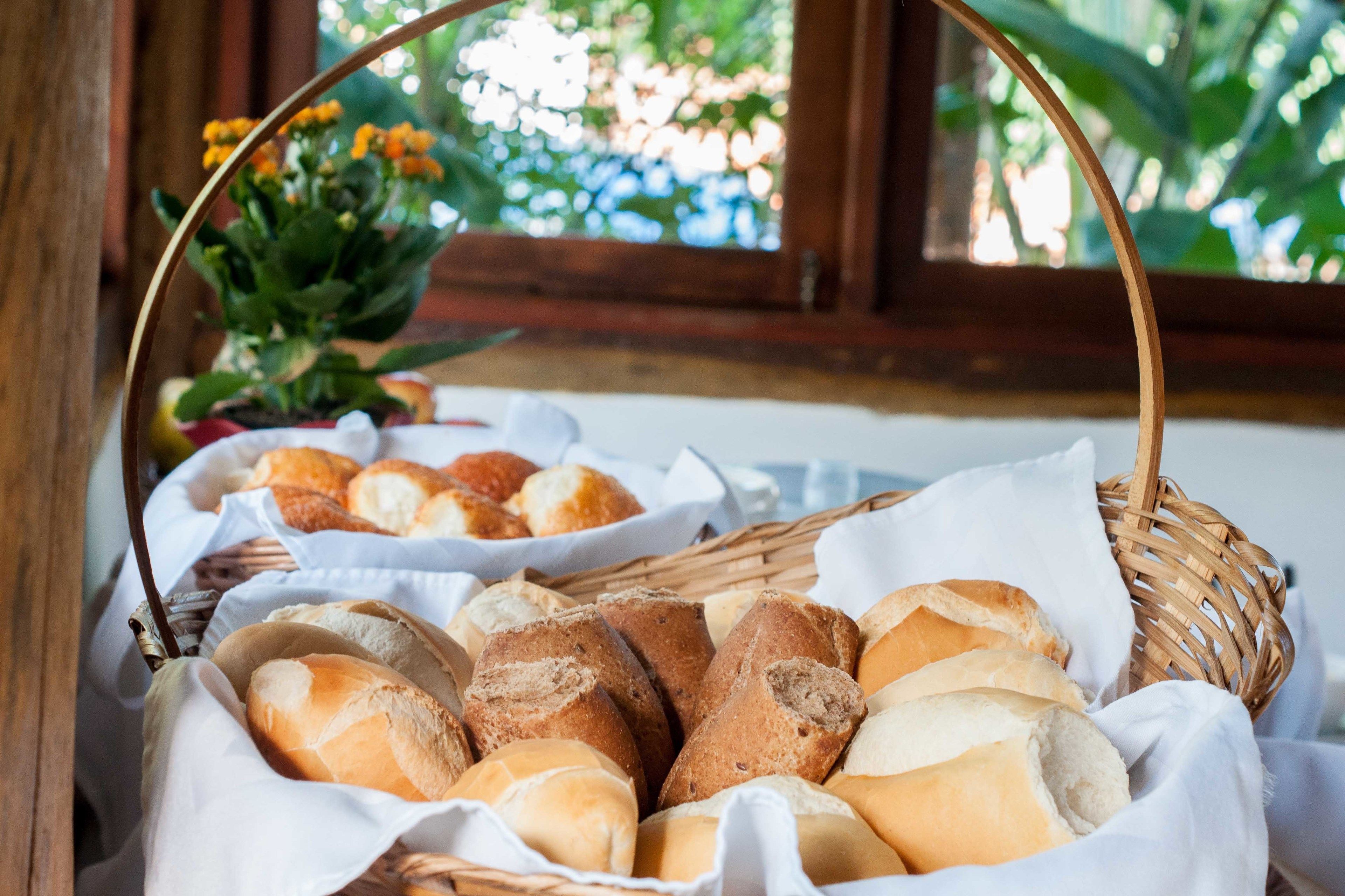 Uma cesta de vime cheia de vários tipos de pão, colocada sobre uma mesa perto de uma janela com vista para o verde.