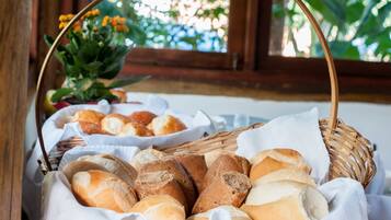 Uma cesta de vime cheia de vários tipos de pão, colocada sobre uma mesa perto de uma janela com vista para o verde.