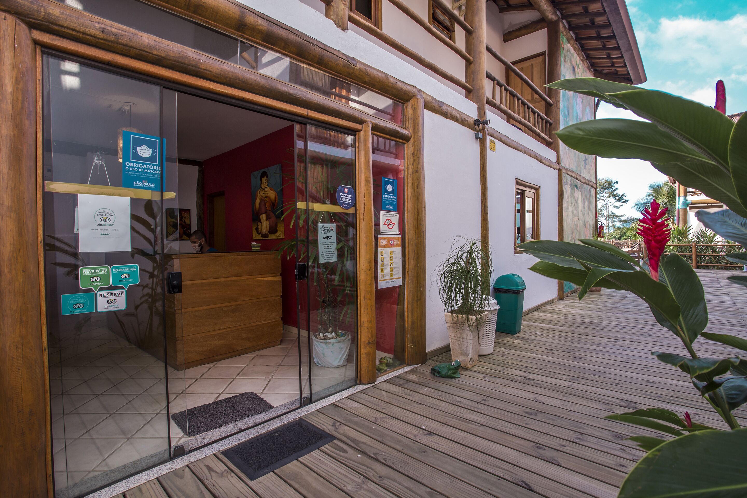 Uma área de recepção com uma mesa de madeira, portas de vidro e diversos avisos, incluindo a obrigatoriedade de uso de máscara.