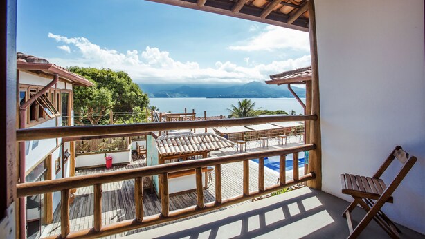 Un balcon doté d’une balustrade en bois, donnant sur un espace piscine avec des chaises longues et des parasols, et offrant une vue sur les montagnes et un lac.