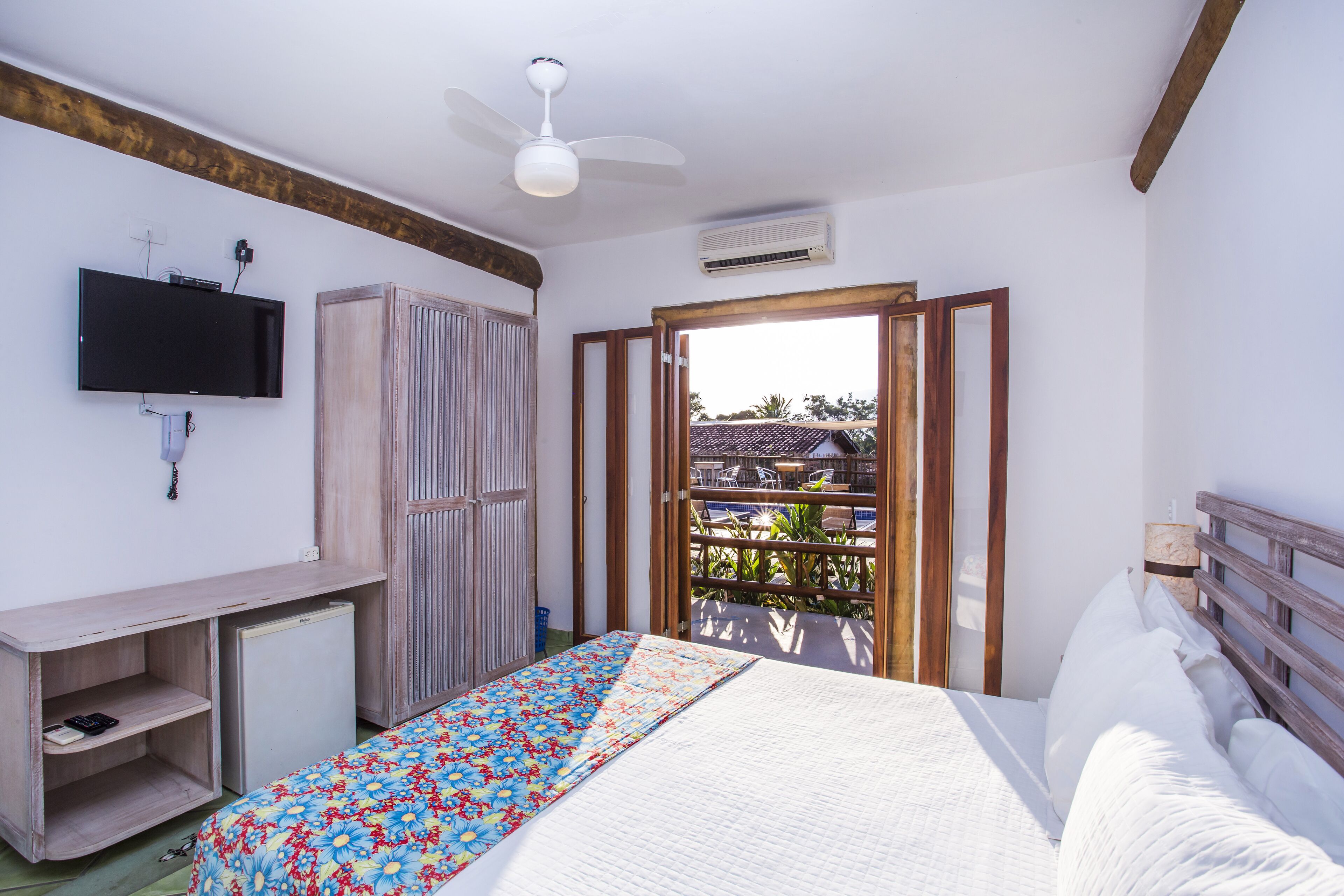 A bedroom with a bed, a TV mounted on the wall, a ceiling fan, and an open door leading to an outdoor area.