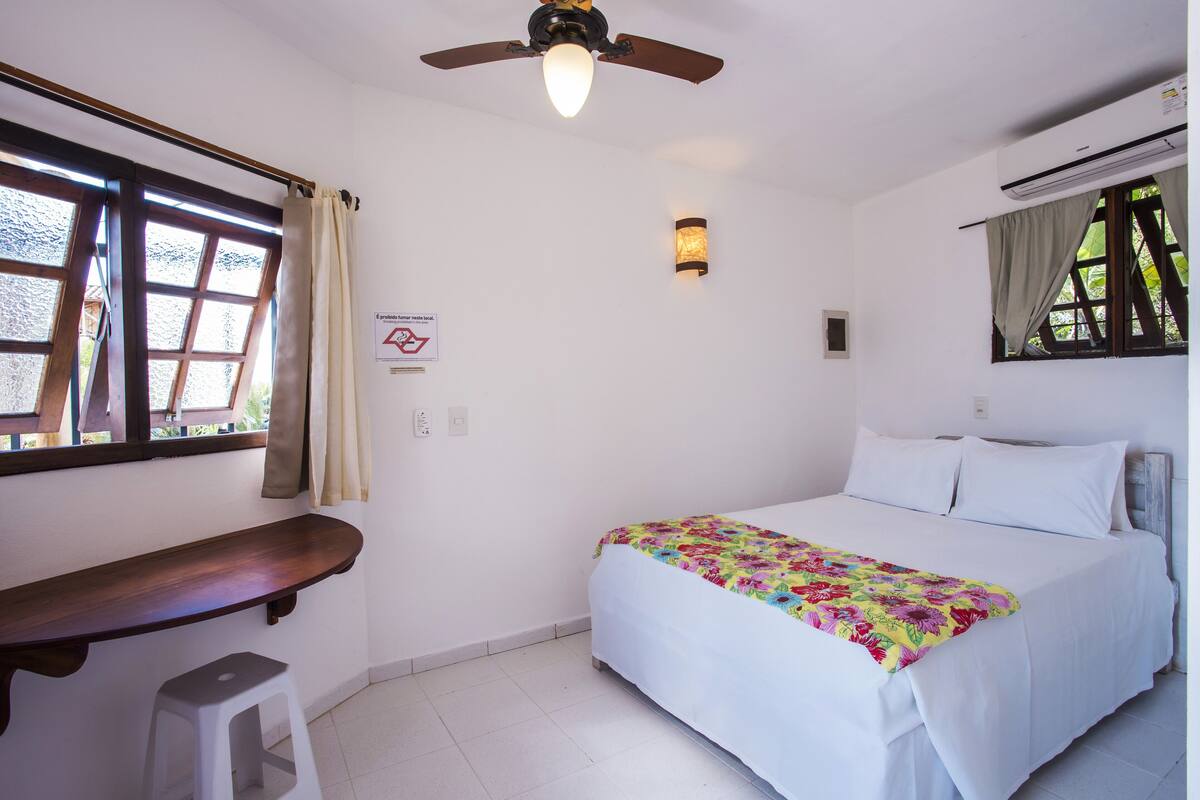 A bedroom with a bed, a desk, a stool, a ceiling fan, a window with curtains, and a wall-mounted light fixture.