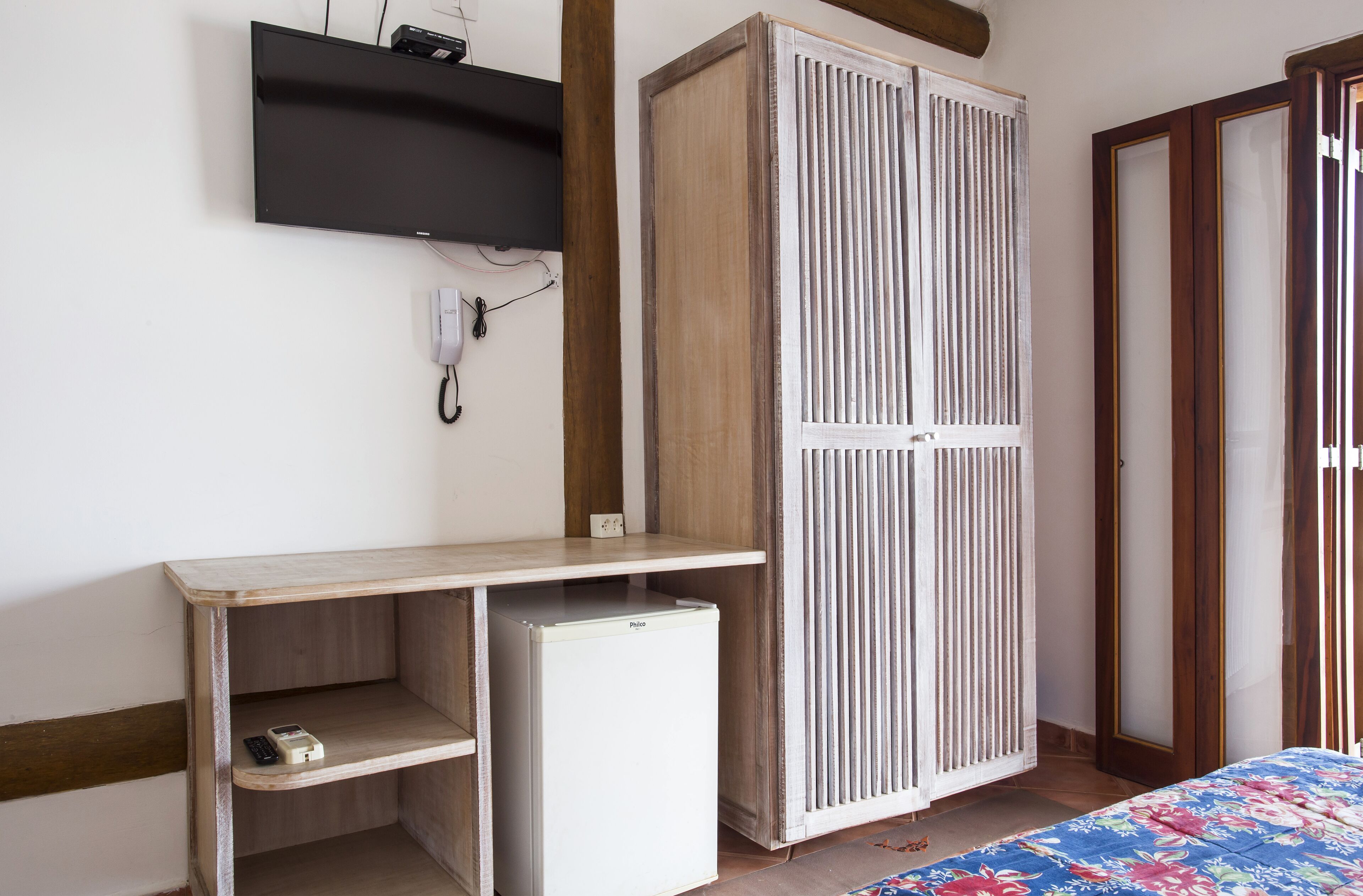A room with a wooden wardrobe, a small white fridge, a desk with a shelf, and a bed with a floral bedspread.