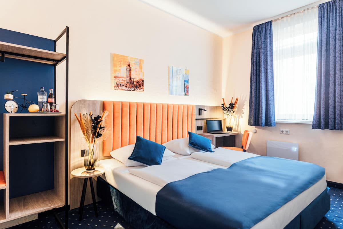 Hotellihuone, jossa on suuri sänky, yöpöytä lampun kanssa, pieni sivupöytä koriste-esineitä varten, kirjahylly ja ikkuna, jossa on verhot.